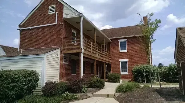 Brick building with a wooden balcony, surrounded by bushes and a small shed. Overcast sky.