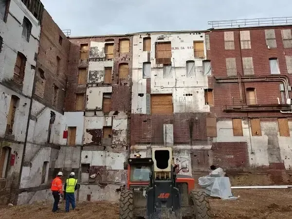 Workers inspect partially demolished brick building with boarded windows. Heavy machinery in foreground.