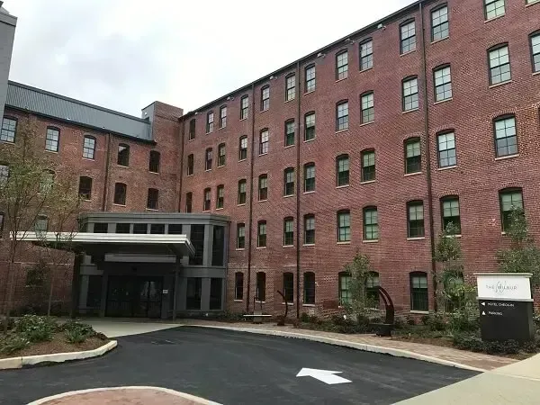 Red brick building with many windows, dark entrance canopy, paved driveway, cloudy sky.