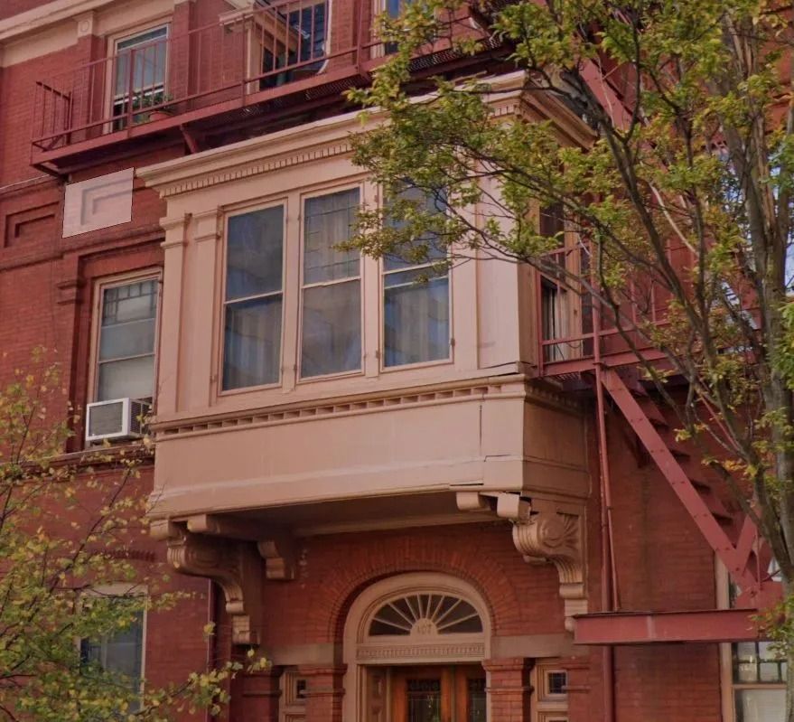 Red brick building with a decorative, windowed bay and fire escape; tree branches frame the right side.