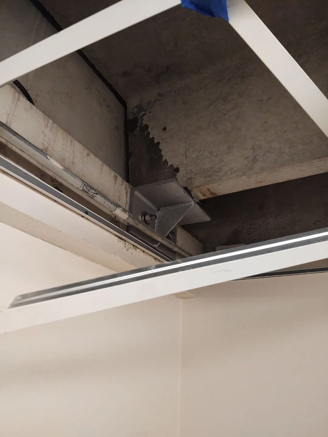 Gray and white suspended ceiling construction with exposed concrete overhead; an angled metal bracket is attached.