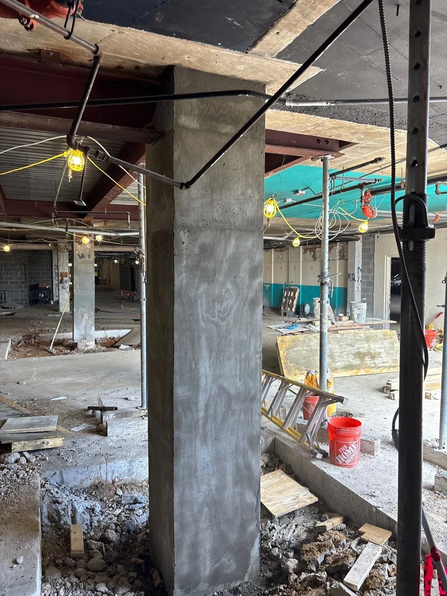 Construction site: concrete column, exposed beams, and debris. Workers and equipment visible in the background.