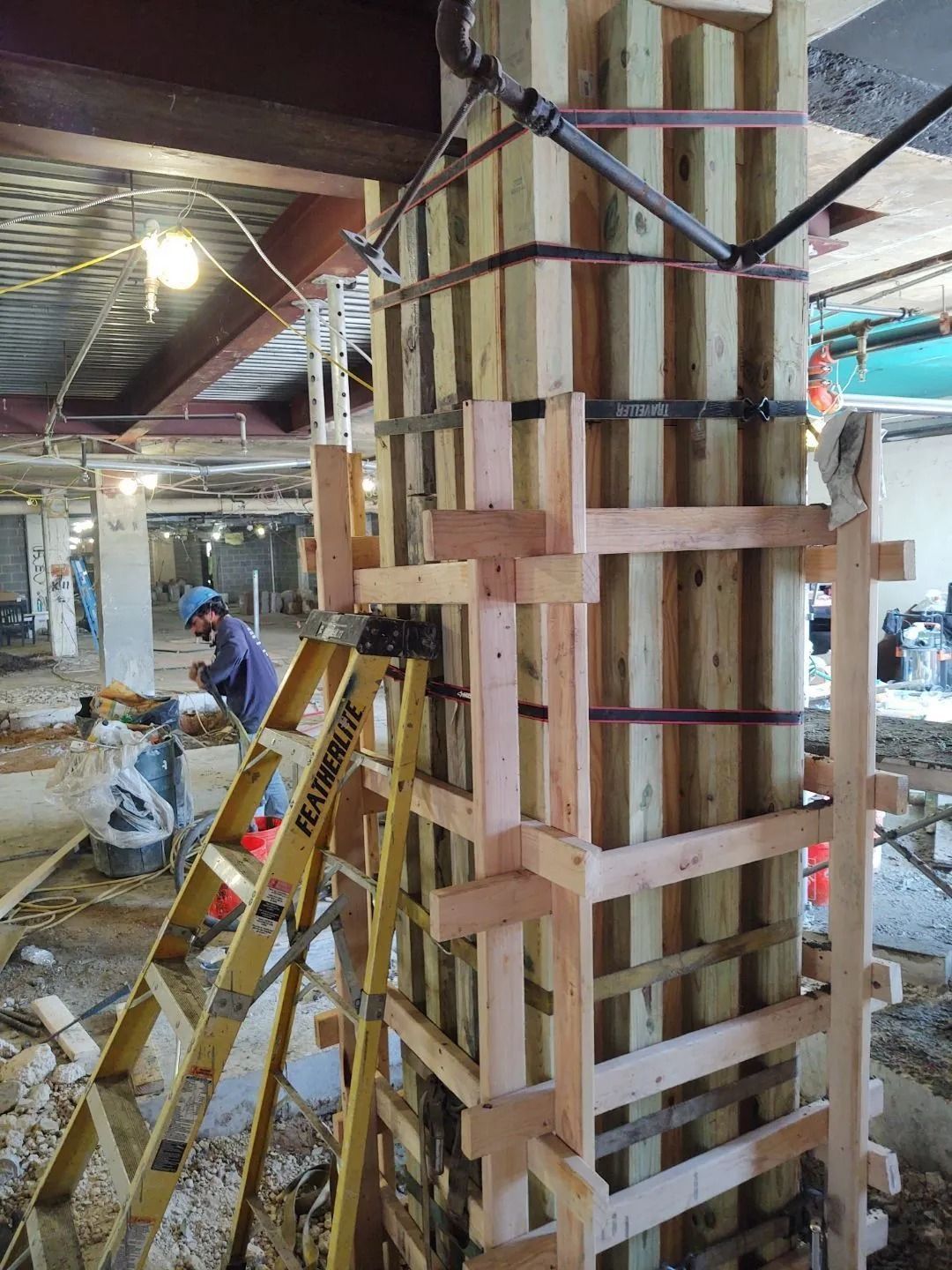 Construction site: Wooden formwork surrounds a structural column. A worker on a ladder, drills.