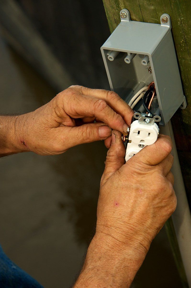 Hands wiring an electrical outlet inside a gray junction box on a wall.