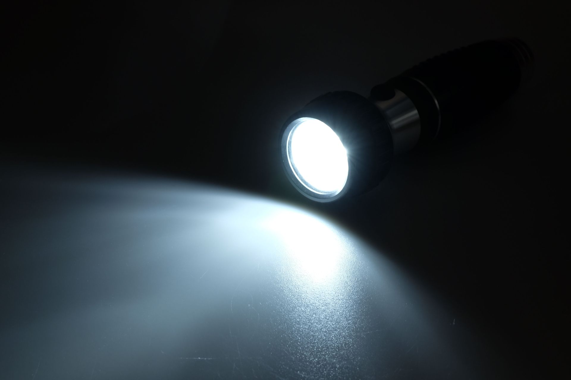 A bright white flashlight beam illuminates a dark surface; the flashlight is black and silver.