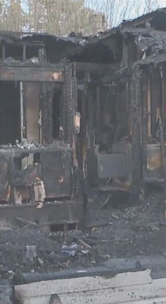 Charred remains of a house after a fire. Blackened wood and debris are visible; the roof is mostly gone.