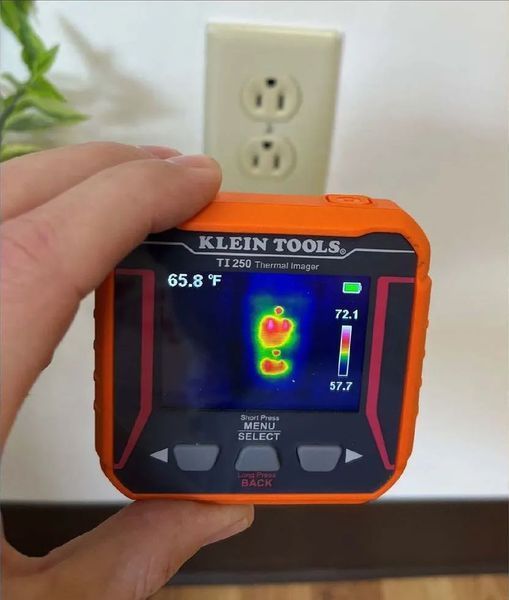 Hand holding thermal imager pointed at electrical outlet, showing heat signature in red and yellow.