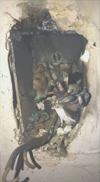Burnt electrical outlet in wall, with charred wires and debris. Black, white, and green discoloration.