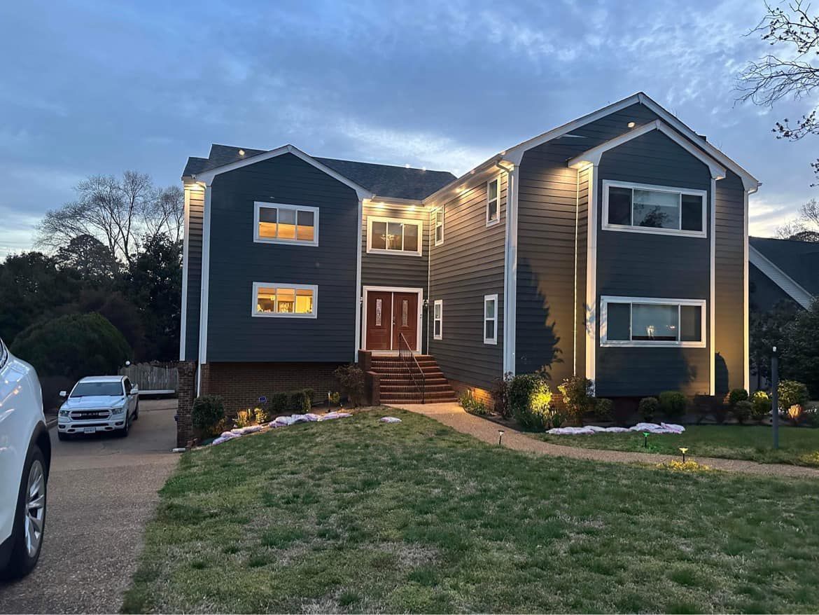 Two-story house with dark gray siding, lit pathways, and a white car in the driveway at dusk.