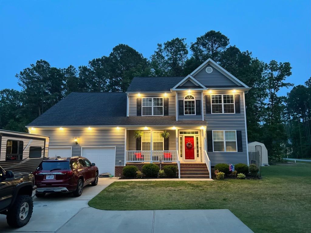 Two-story house with blue-gray siding, black shutters, lit porch, red front door, and parked vehicles at dusk.