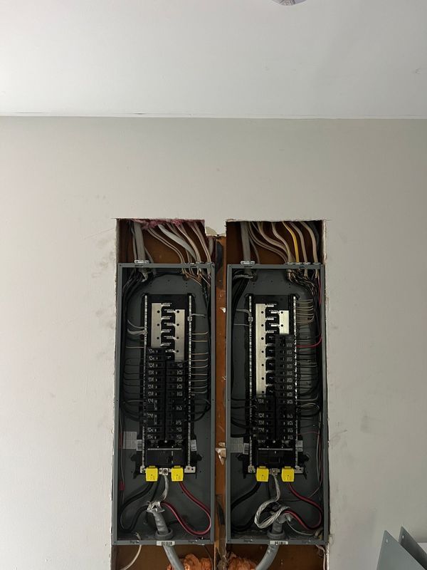Two electrical breaker panels installed in a wall opening, with exposed wires.