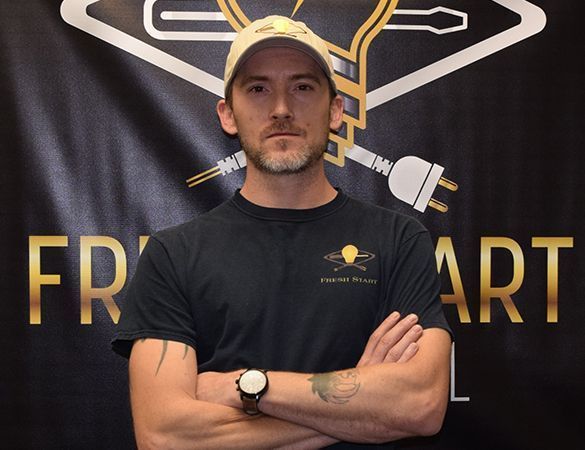 Man with arms crossed, wearing a hat and t-shirt, standing in front of a logo with a lightbulb and plug.