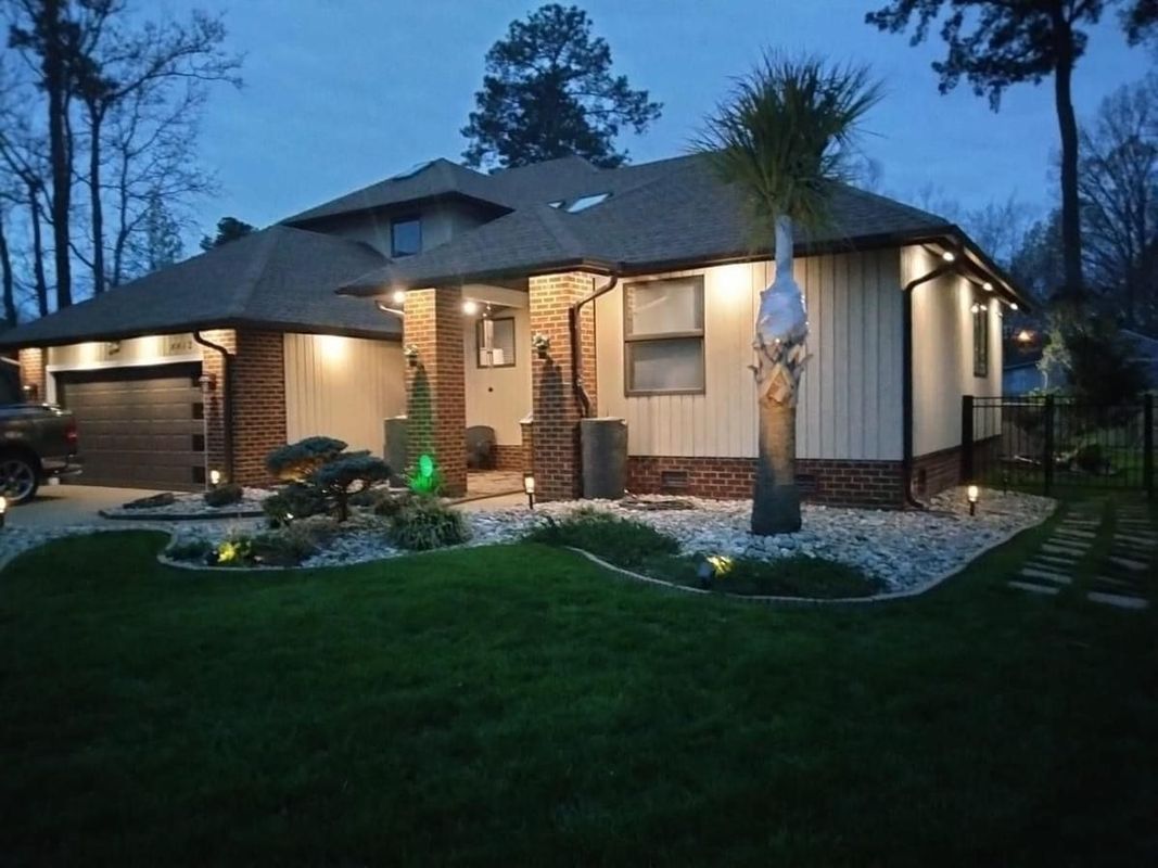 House exterior at dusk, with lit landscape and spotlights. Features brick and light siding, with a palm tree.