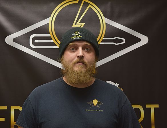 Man with beard wearing beanie and navy t-shirt, standing in front of a logo.