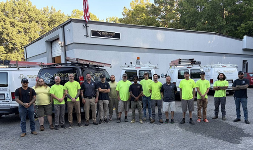 Group of workers in neon shirts standing in front of utility trucks and a building during daylight.