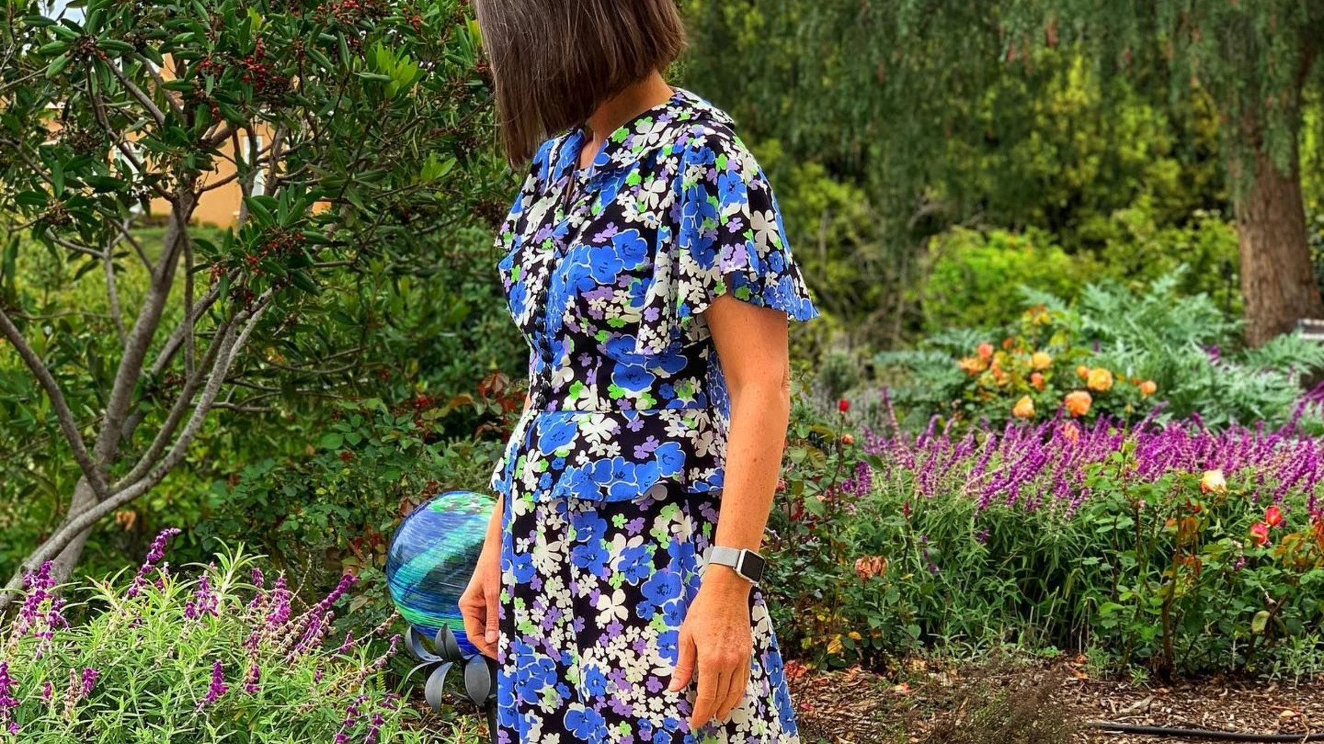 Woman in blue floral dress stands in a garden.