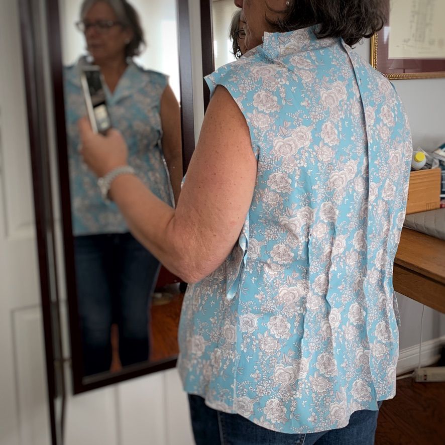 Woman in blue floral top, standing near a mirror, holding a phone, taking a photo.
