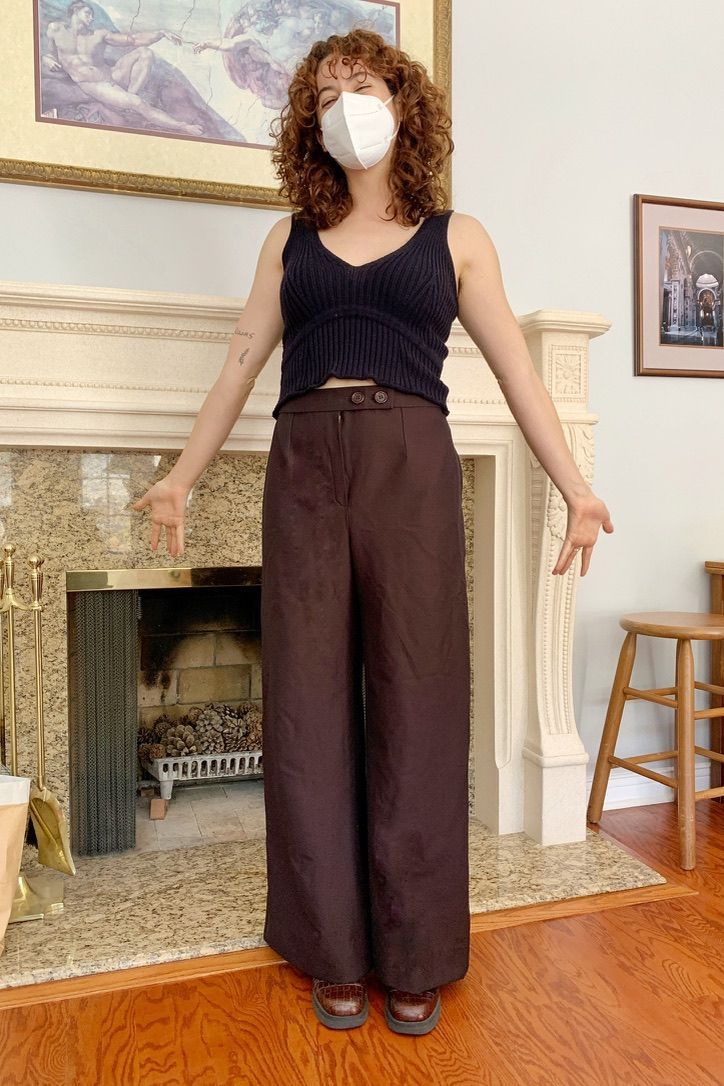 Person wearing a mask, navy top, and brown pants, standing in front of a fireplace with arms outstretched.