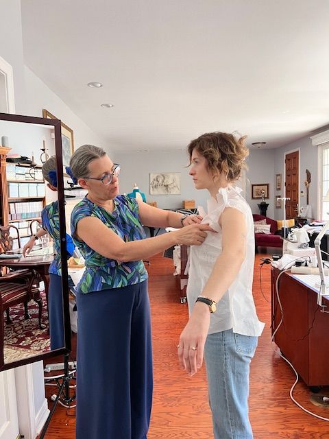 Woman in blue floral top adjusts a white top on another woman in front of a mirror in a well-lit room.