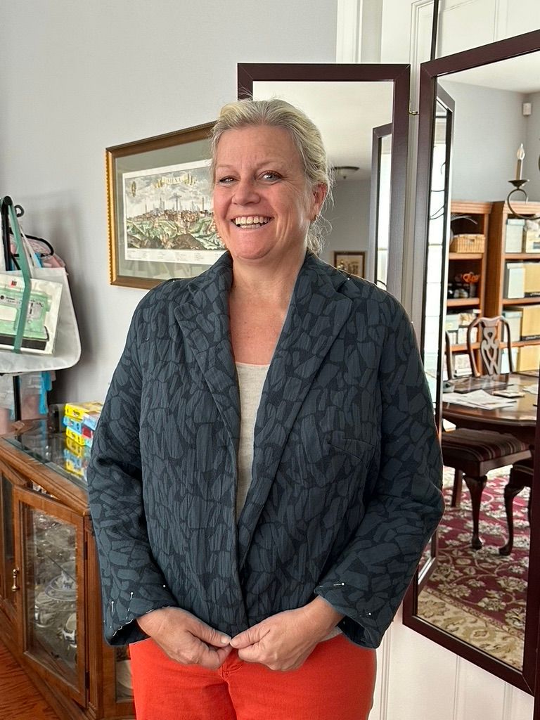 Woman in a dark patterned jacket and orange pants smiles, standing indoors near a mirror.