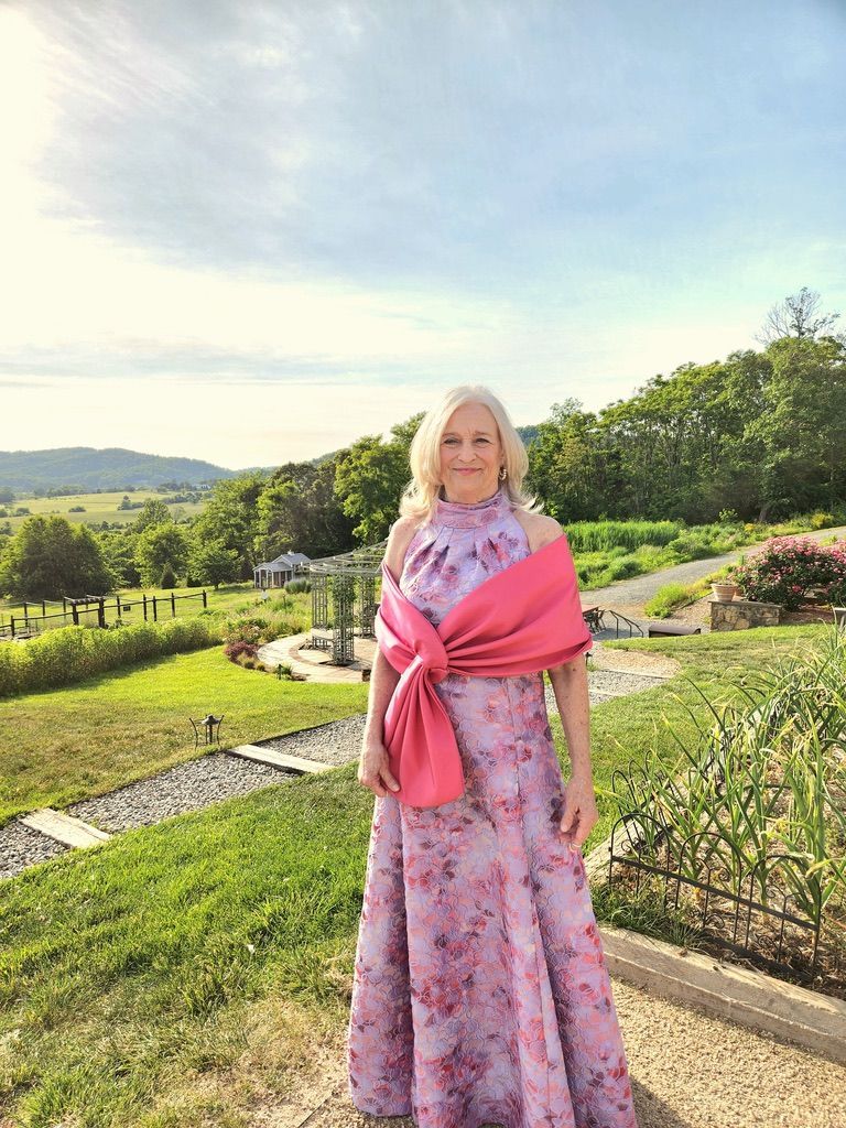 Woman in a pink floral gown and shawl, standing outdoors with a scenic background of hills and a garden.