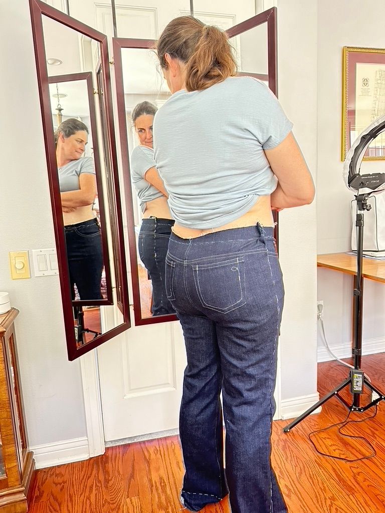 Woman trying on dark blue jeans in front of a three-panel mirror, in a room with a light and wooden furniture.