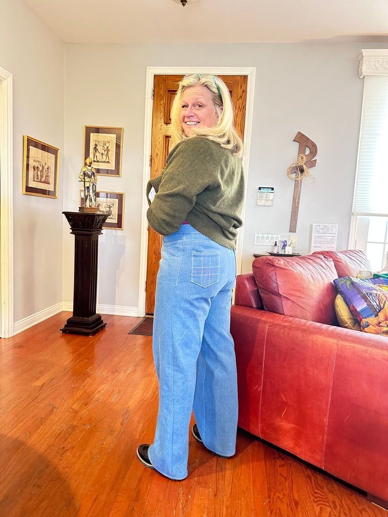 Woman in green sweater and high-waisted, wide-leg blue jeans stands indoors, facing away, smiling. Wooden floor.