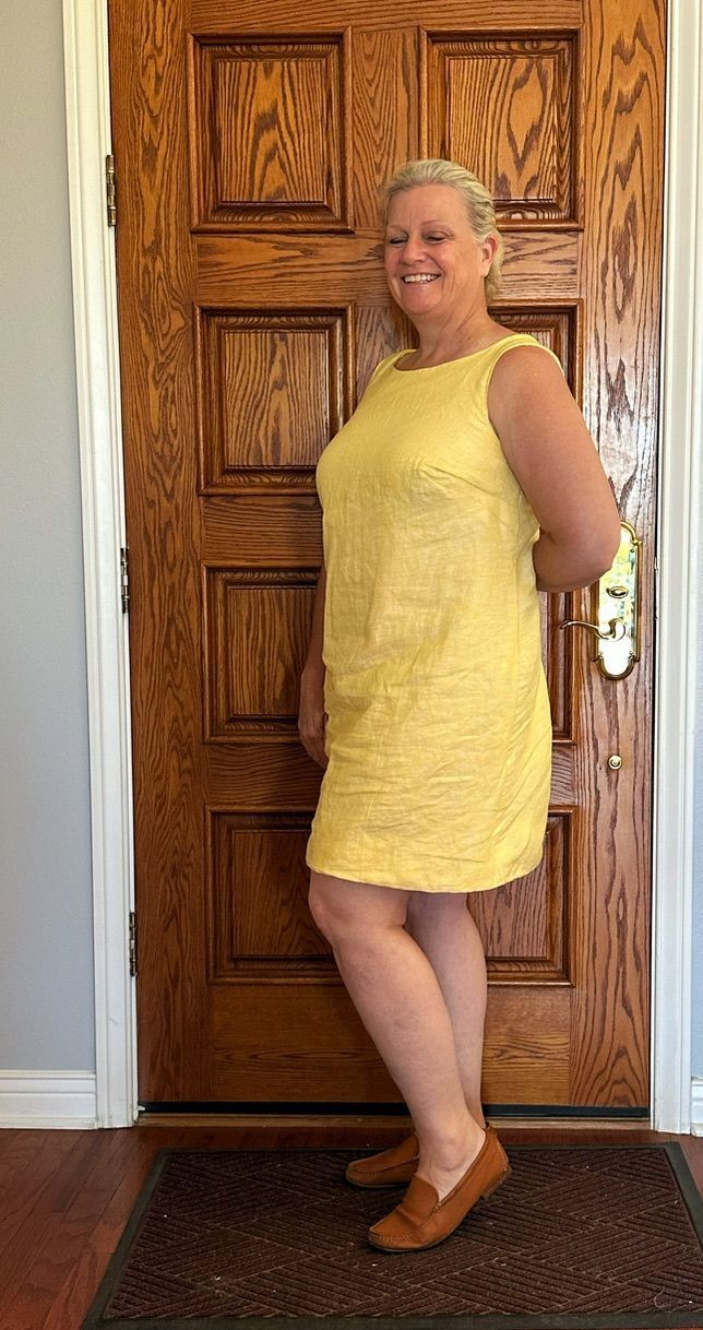 Woman in a yellow dress stands by a wooden door, smiling, wearing brown loafers.