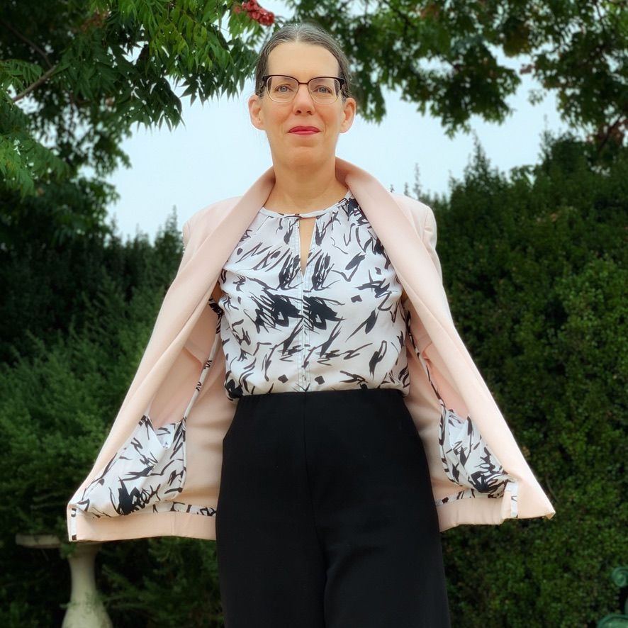 Woman in pink jacket over black pants and patterned top, standing outside.