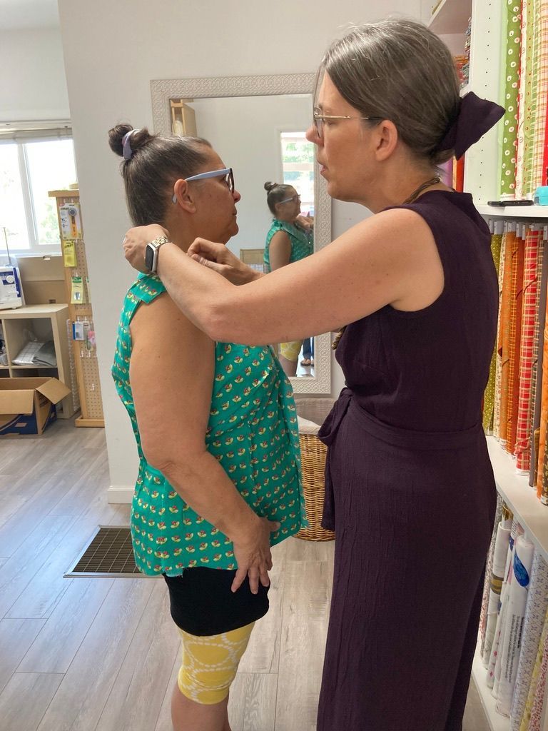 Woman in purple dress measuring another woman's shoulder, fabric store interior.