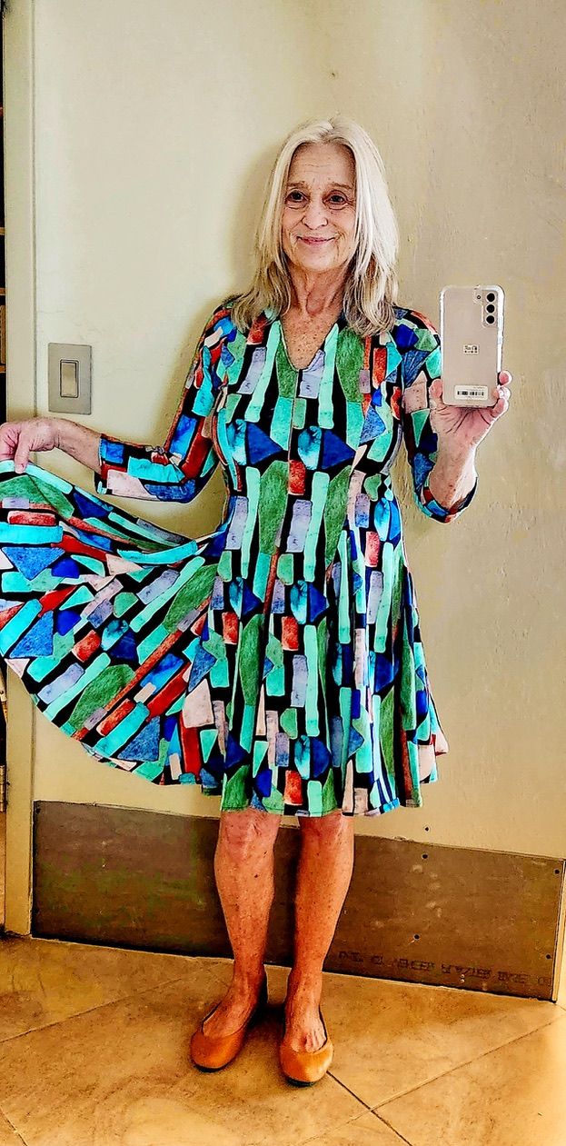 Woman in a patterned dress and ballet flats holds a phone, smiling. Indoors, dress flared.