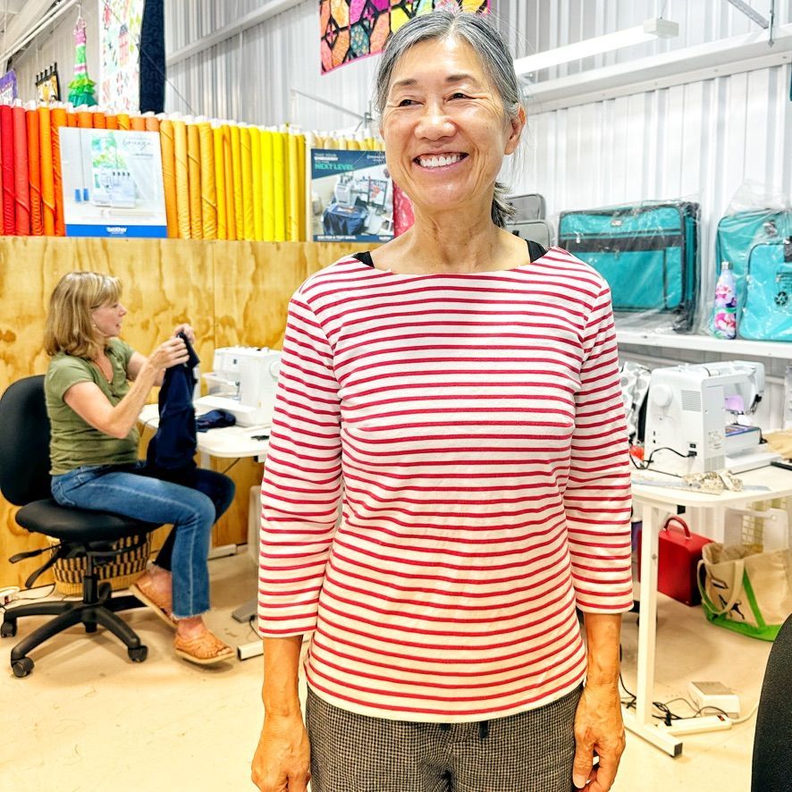Woman in red striped shirt smiles in a sewing workshop. Another woman sews at a table behind her.