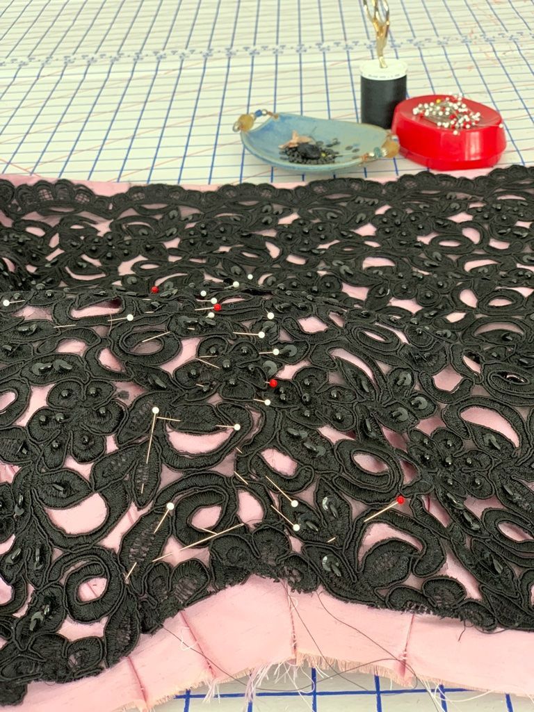 Black lace fabric pinned to pink fabric on a grid surface, sewing notions visible.