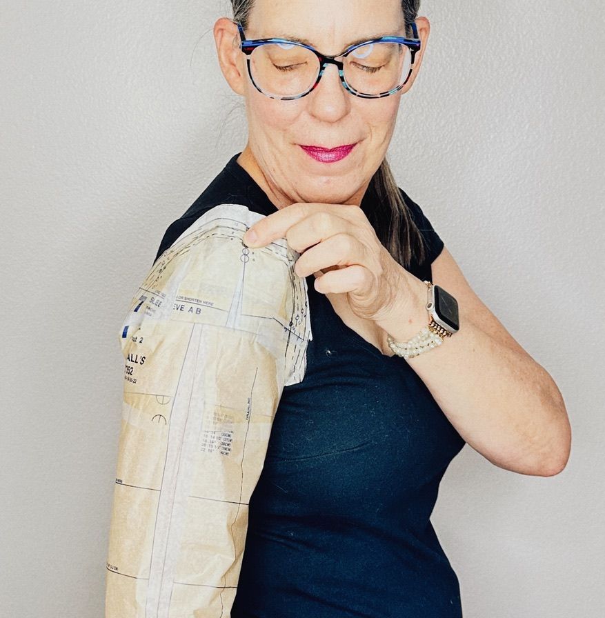 Woman holds a pattern over her arm, comparing fit. She wears glasses and a black shirt.
