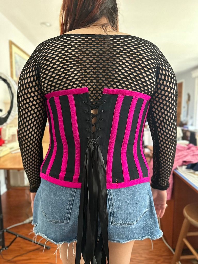 Woman wearing a black and pink striped corset over a black fishnet top and denim shorts, back view.