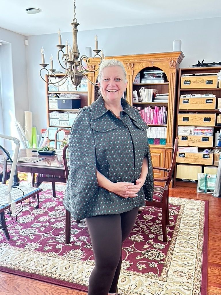 Woman in patterned cape, standing in front of a bookcase, table, and chandelier in a room with a patterned rug.