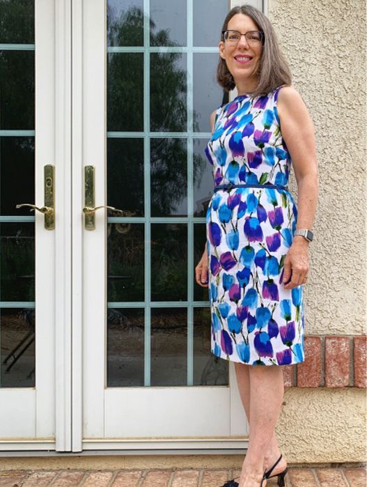 Woman in a blue and purple patterned dress standing by glass doors and a stucco wall.