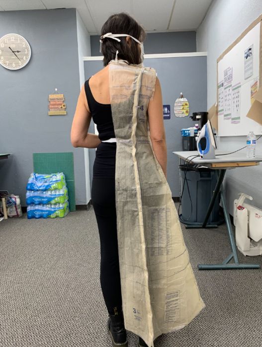 Person in a black outfit with a long beige fabric train, standing in a clinic room with a wall clock and exam table.