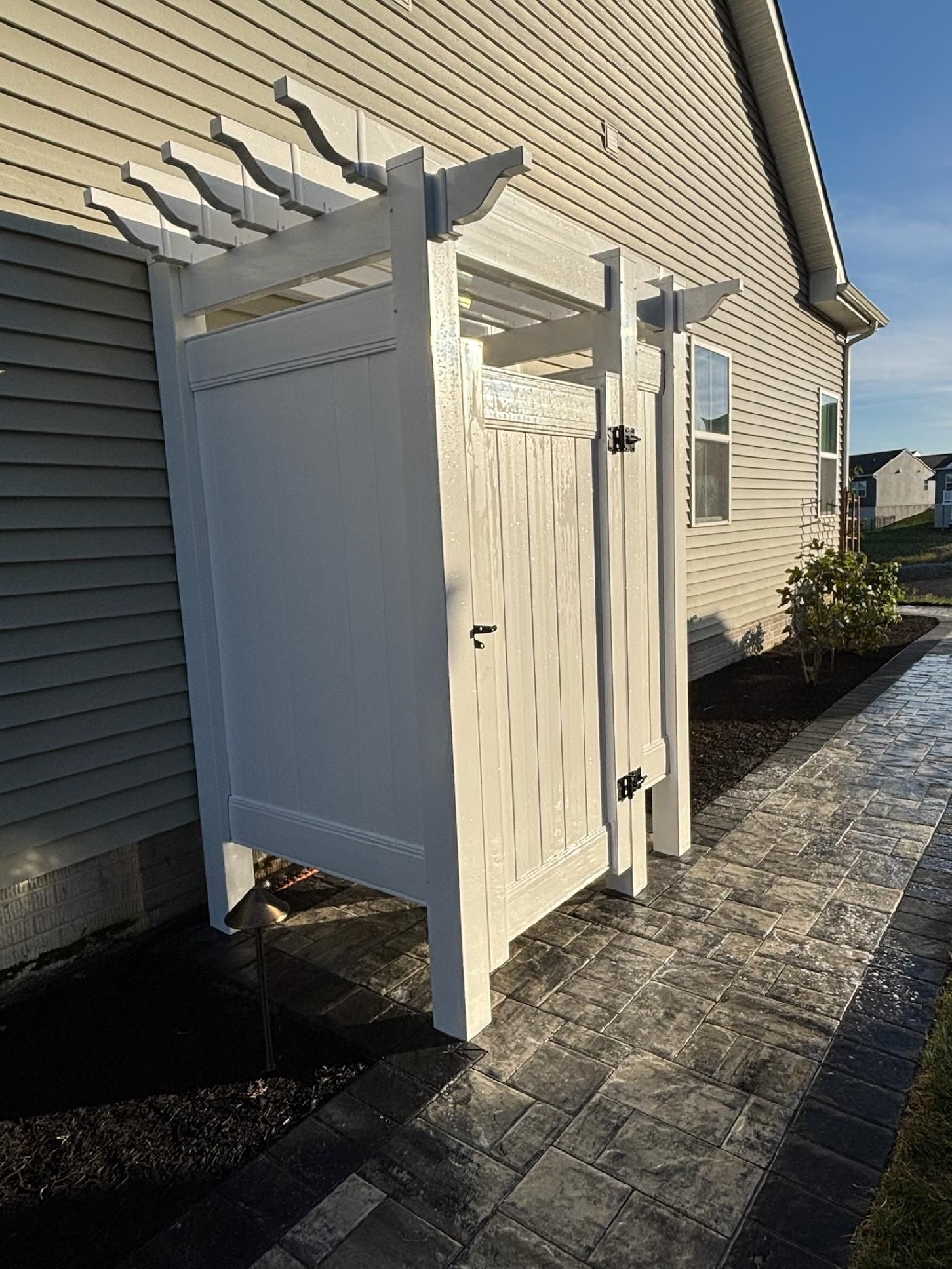 White outdoor shower against a light gray house; a walkway runs alongside the shower.