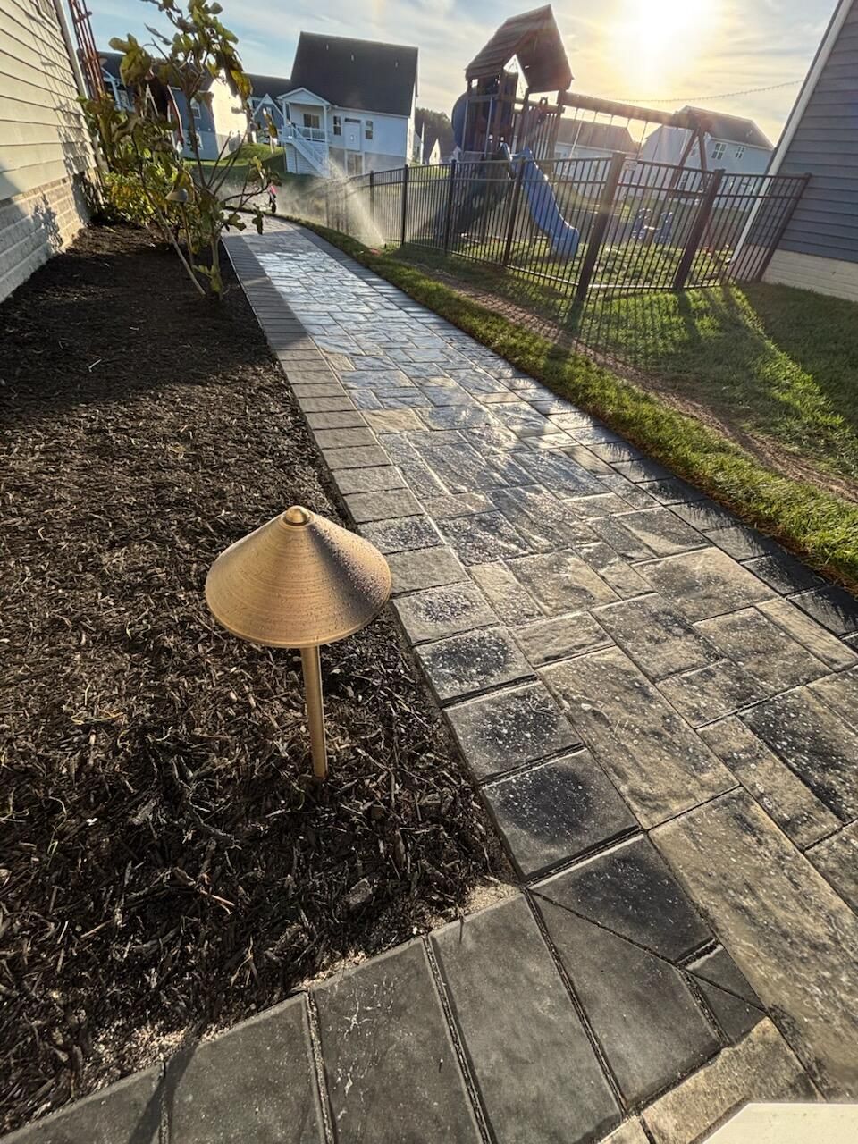 Brick pathway with a mushroom-shaped light, bordered by mulch and grass; a swing set in the background.