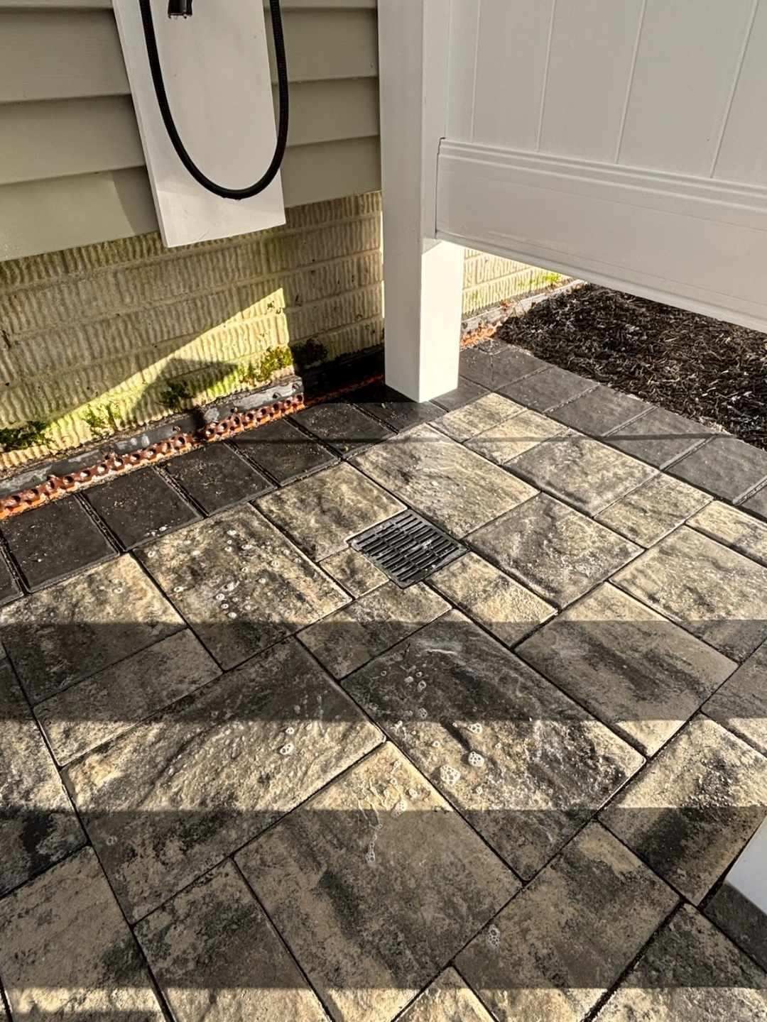Brick patio with a drain, next to a white pillar, with a house in the background.