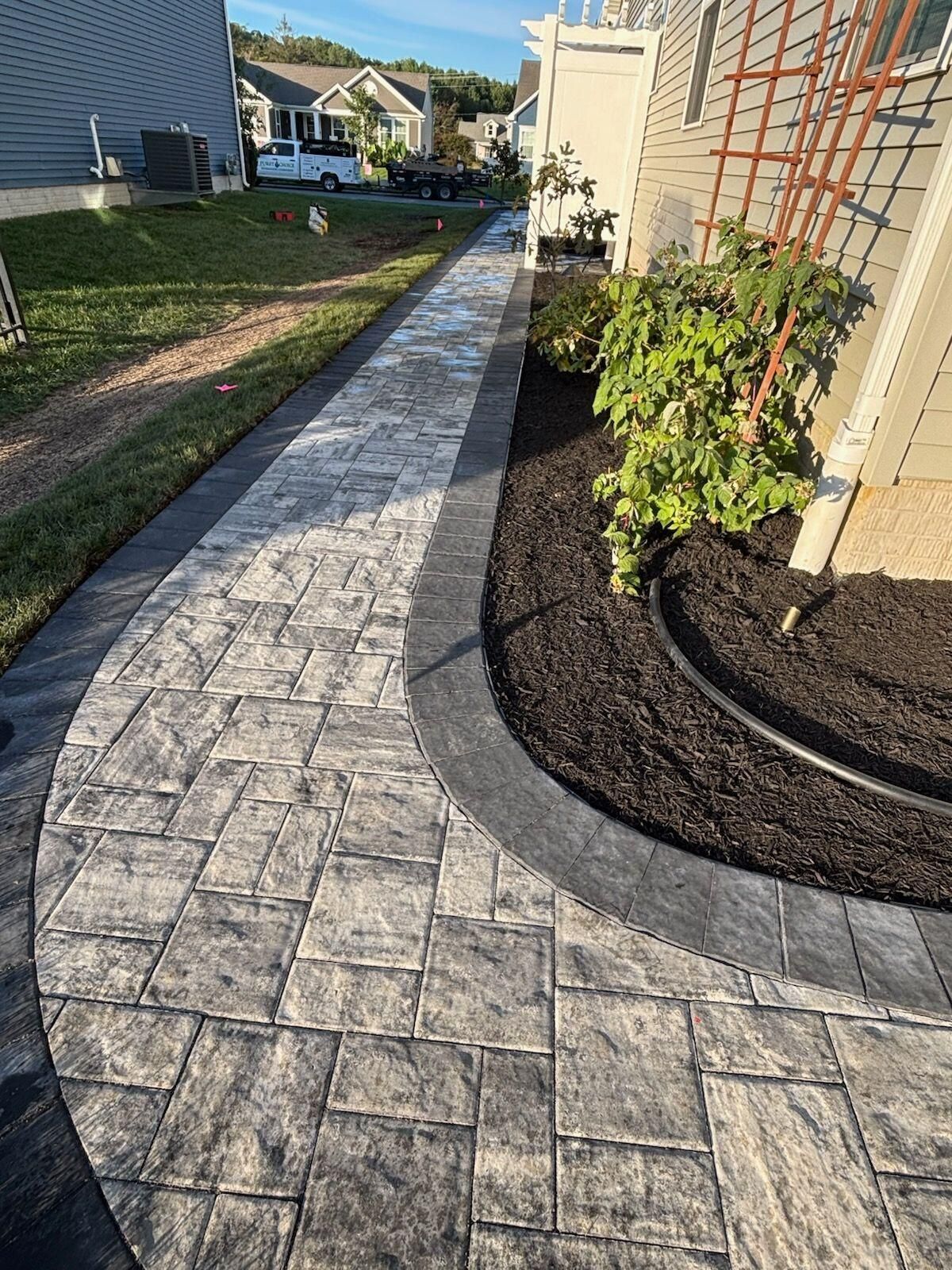 Brick walkway curving past a house with landscaping. Gray pavers edged with dark borders.