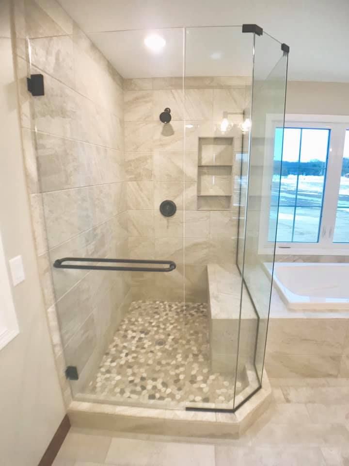 Modern glass walk-in shower with beige tile, grab bar, built-in shelf, pebble floor, and a window nearby