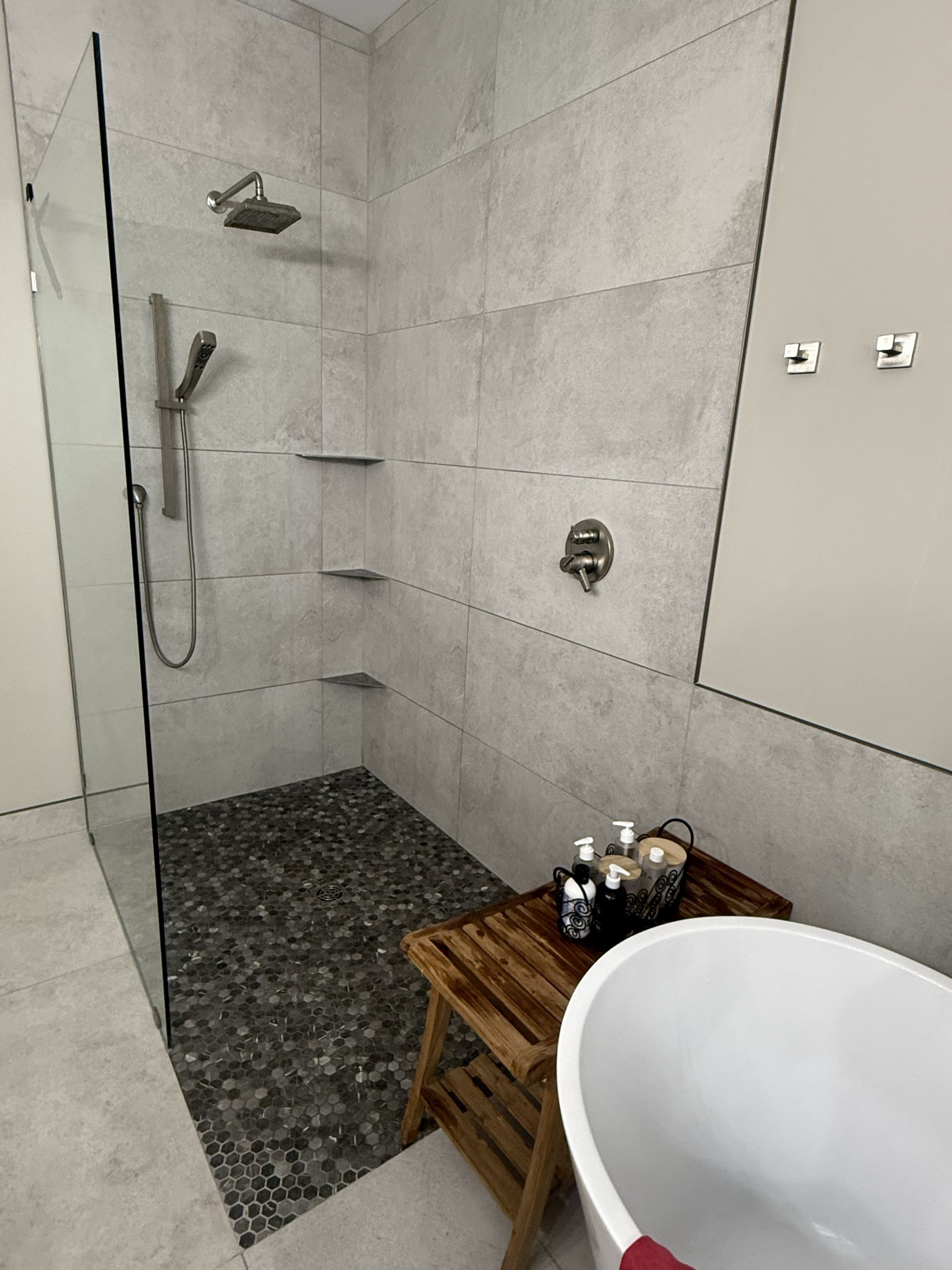 Modern walk-in shower with gray tile, black floor, wooden stool, and white bathtub edge