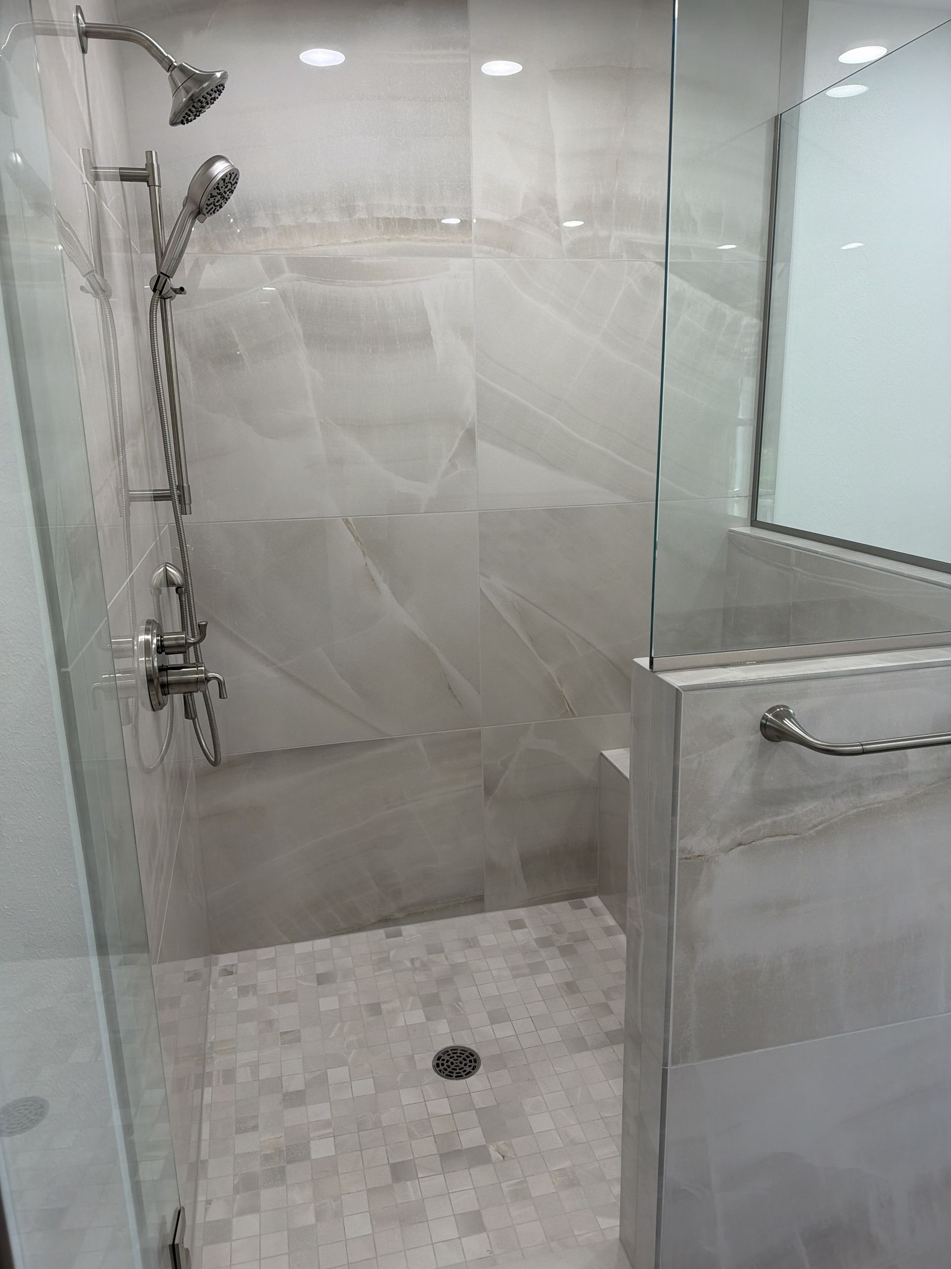 Accessible tiled shower with grab bars, handheld showerhead, and built-in bench beside a glass partition