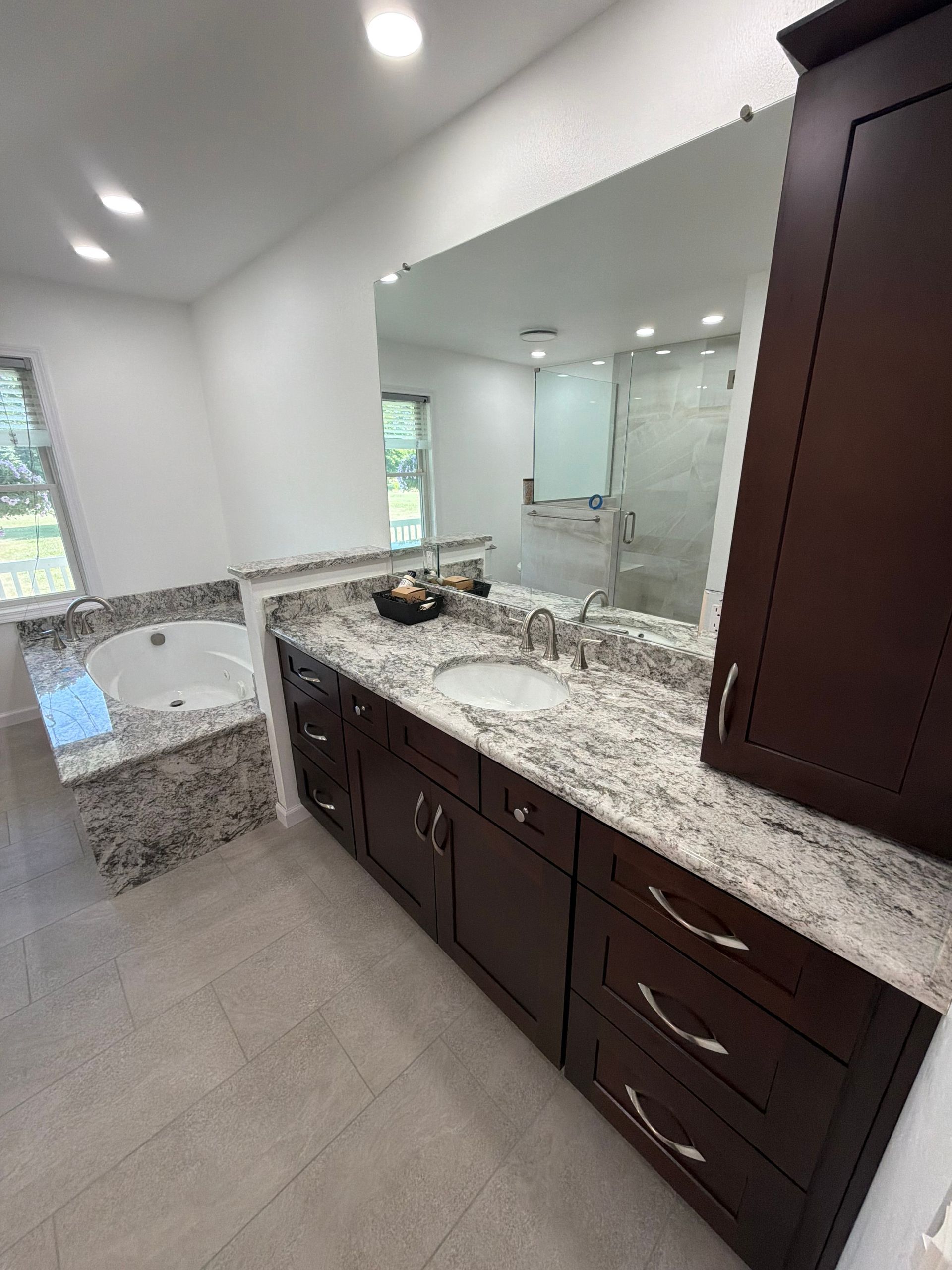 Modern bathroom vanity with dark wood cabinets, granite countertop, mirror, and adjacent soaking tub.