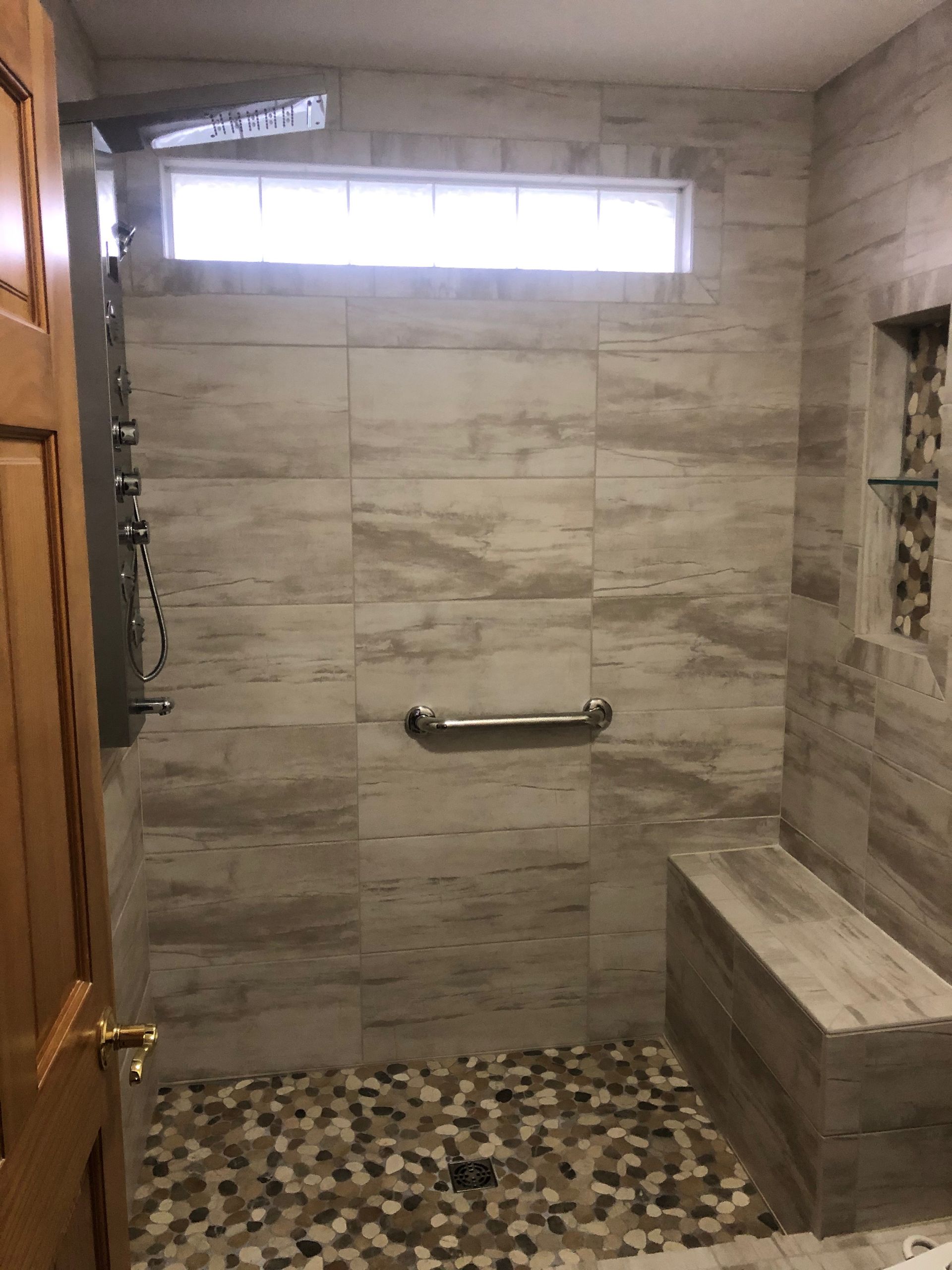 Accessible shower with gray tile walls, pebble floor, grab bar, bench, and a small high window.