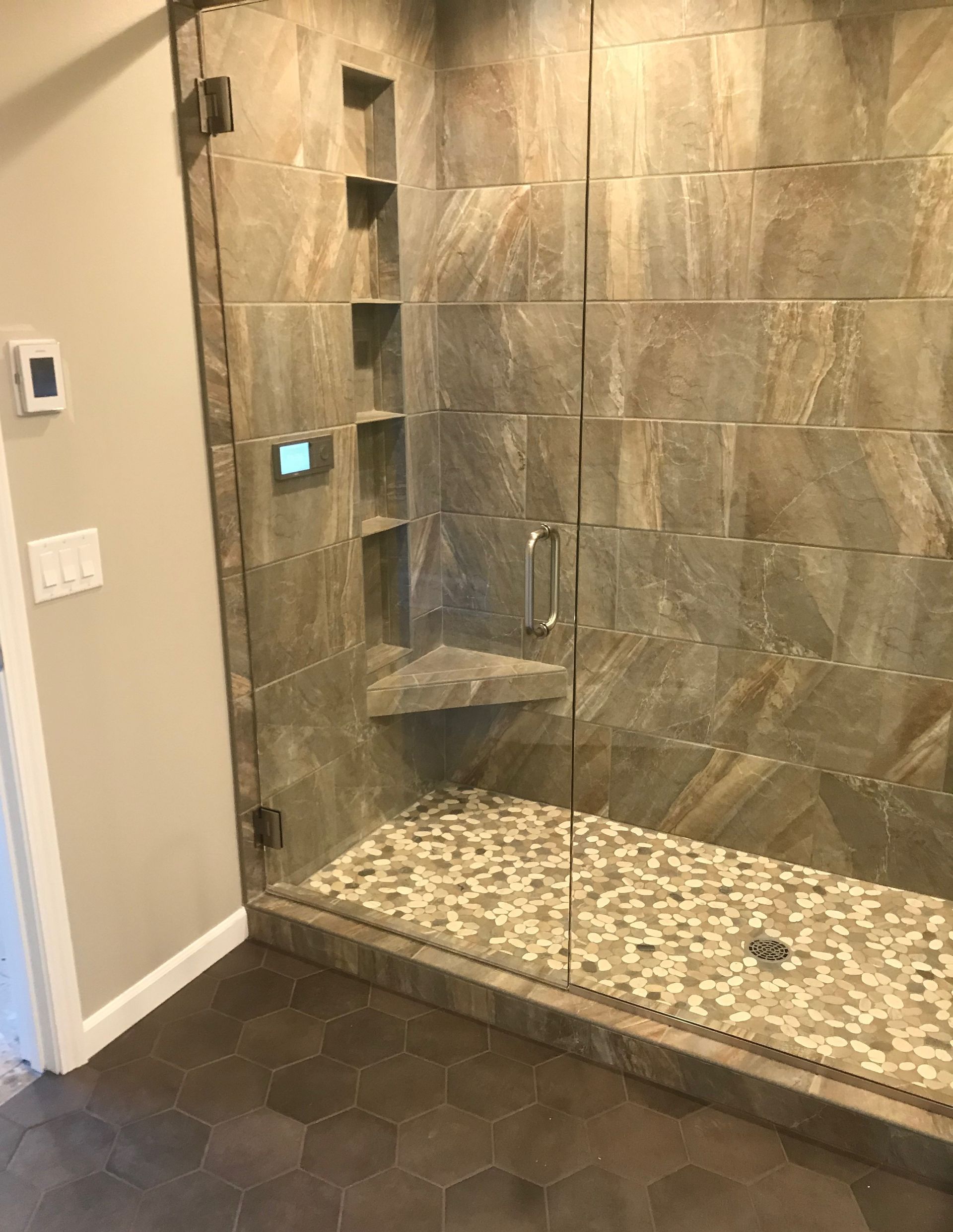 Glass walk-in shower with stone-tile walls, pebble floor, and dark hexagonal bathroom tile outside.