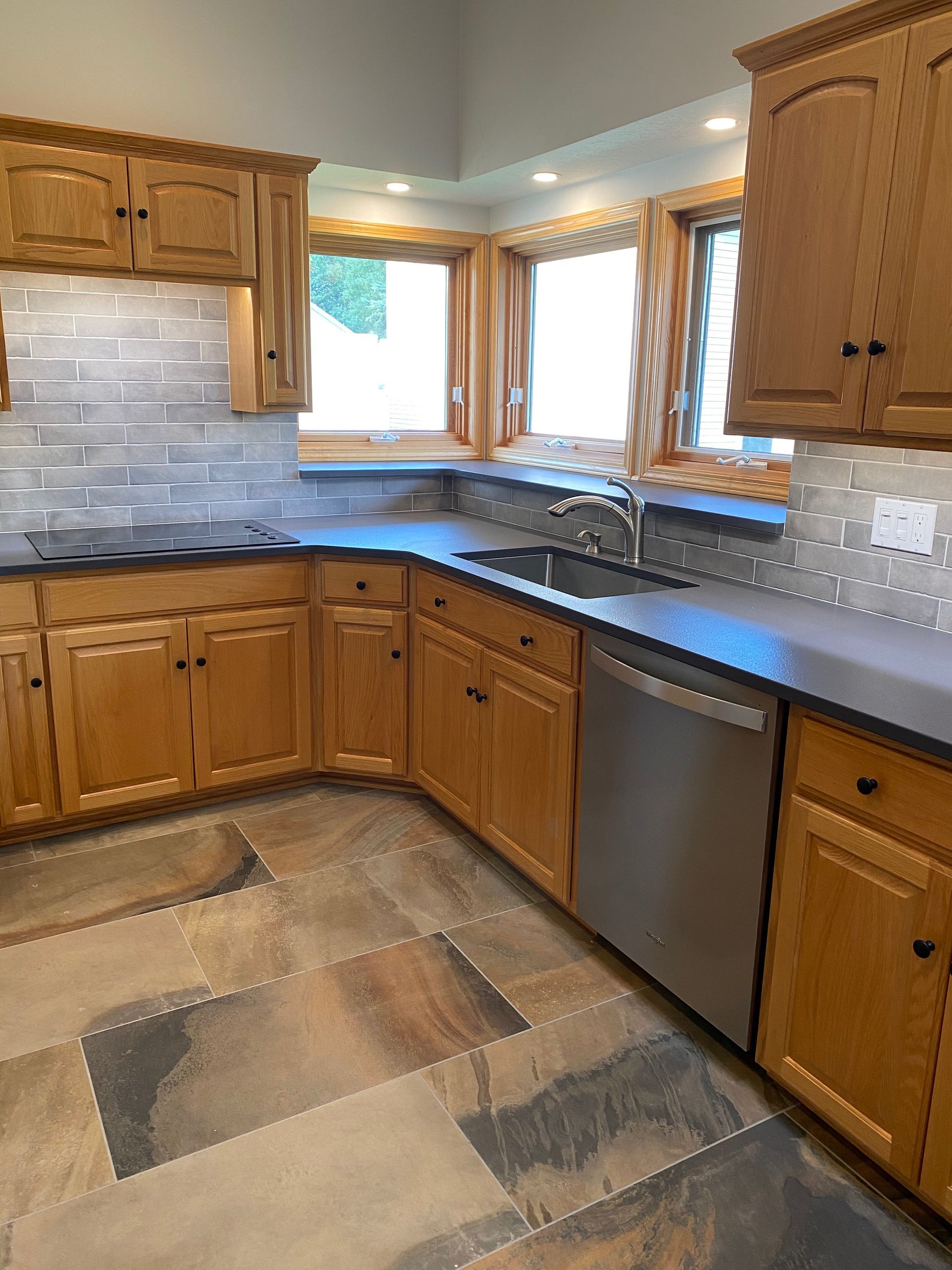 Wooden kitchen with gray countertops, tiled backsplash, corner windows, and stainless steel dishwasher.