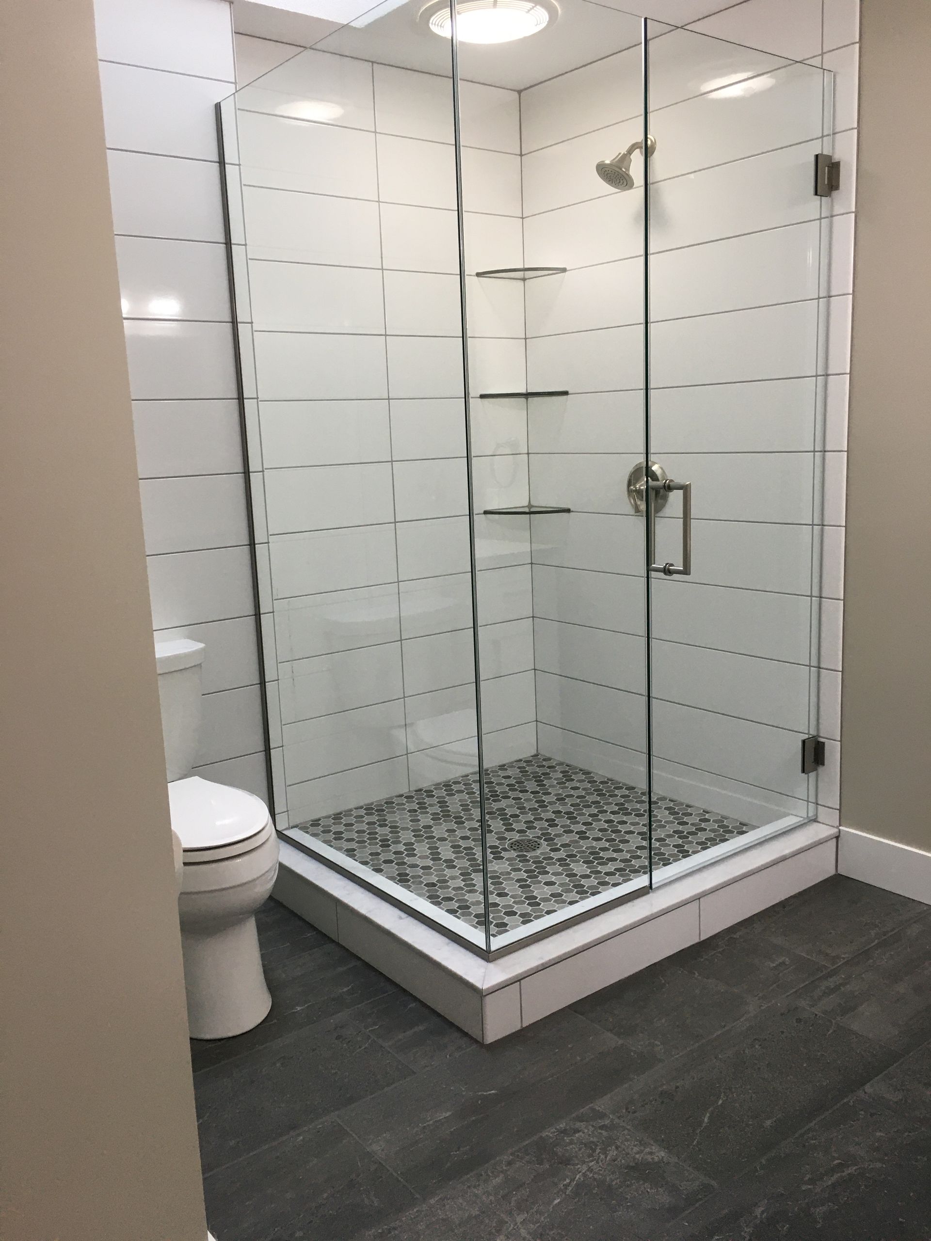 Glass shower stall in a modern bathroom with white tile and dark gray floor.